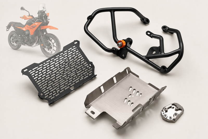 Adventure motorcycle protection accessories including a black tubular crash guard, radiator grill, aluminium bash plate and side stand base on a bone white studio background